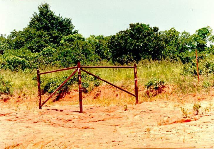 An intersection of two country roads, where erosion has stripped away the soil and almost unearthed the fenceposts, anchored in the Permian