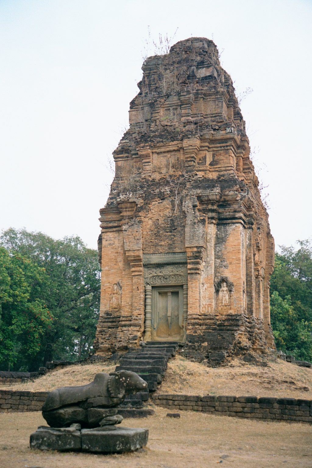 Bakong's pyramid is ringed with eight brick towers, comparable to those at Preah Ko. In front of this one, on the west, sits Nandi, Shiva's